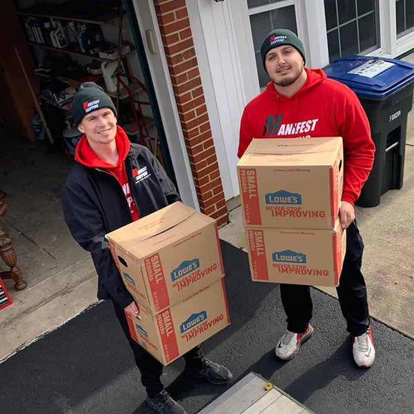 two manifest shipping movers with branded hats and hoodies, holding a pair of moving boxes in their hands