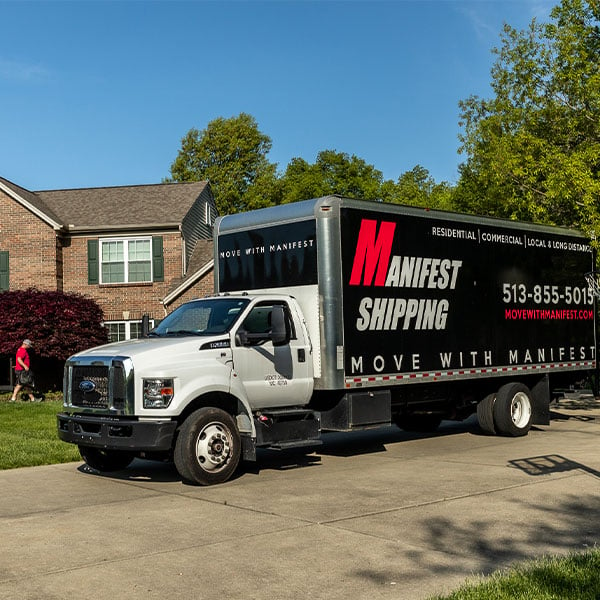 we see manifest shipping moving truck with company details on it in the foreground, in the backroundd, there is a mover wearing red shirt preparing for the move in front of the house